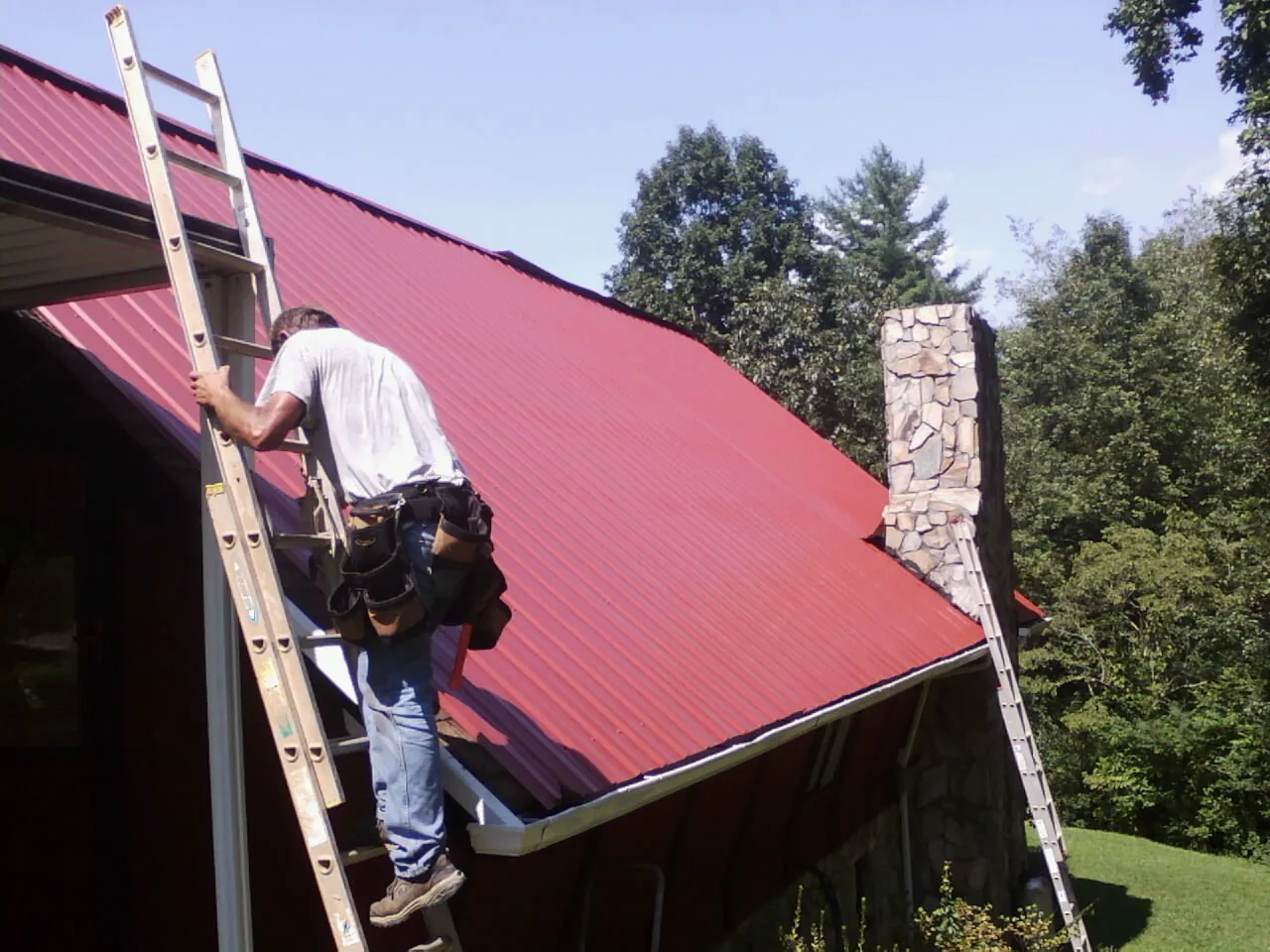 Roofer on a ladder beside a red metal roof and stone chimney — Soffit & Fascia Repair by Roofing Brothers in East Syracuse